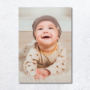 Pu&ograve; includere: Un bambino con un cappello a righe marroni e bianche e un body con motivo beige e marrone &egrave; sdraiato a pancia in gi&ugrave; su una superficie bianca e soffice. Il bambino sorride e guarda la telecamera.