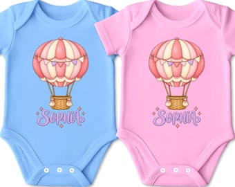 Könnte beinhalten: Zwei kurzärmelige Babybodys, einer in Hellblau und einer in Hellrosa, jeweils mit einer zentralen Grafik eines rosafarbenen und cremefarbenen Heißluftballons mit braunem Korb und Wimpeln, wobei der Name Sophia darunter in Lila gedruckt ist.