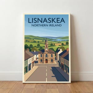 May include: A framed art print featuring a stylized illustration of Lisnaskea, Northern Ireland. The artwork depicts a town square with a clock tower building, surrounded by buildings and a road. Green hills and a blue sky complete the scene.