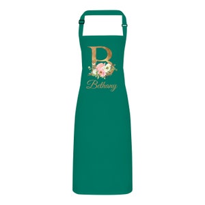 Op de afbeelding: Een groene schort met een gouden letter "B" en een bloemenontwerp. De naam "Bethany" staat in gouden letters onder de letter.