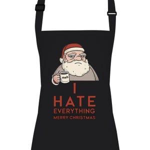 Puede incluir: Delantal negro con una ilustración de un Papá Noel gruñón sosteniendo una taza con la palabra "meh". El texto "I HATE EVERYTHING MERRY CHRISTMAS" está impreso en rojo. El delantal tiene una correa para el cuello y lazos negros.