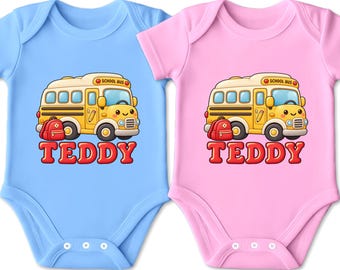 Könnte beinhalten: Zwei kurzärmelige Babybodys, einer in Hellblau und einer in Hellrosa, jeweils mit einer Grafik eines gelben Schulbusses mit rotem Rucksack und dem Schriftzug "TEDDY" in roten Buchstaben unter dem Bus. Die Bodys haben einen Rundhalsausschnitt und Druckknöpfe.