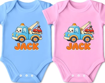 Könnte beinhalten: Zwei kurzärmelige Babybodys, einer in Hellblau und einer in Hellrosa, jeweils mit einer Cartoon-Illustration eines blauen Abschleppwagens, der ein kleines rotes Auto trägt. Unter der Grafik ist der Name JACK in orangefarbenen Buchstaben.
