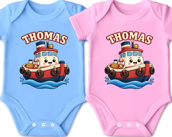 Könnte beinhalten: Zwei kurzärmelige Babybodys, einer in Hellblau und einer in Hellrosa, jeweils mit einer Grafik eines lächelnden rot-weißen Schleppers auf blauen Wellen und dem Namen THOMAS in einer fetten, geschwungenen Schrift über dem Boot.