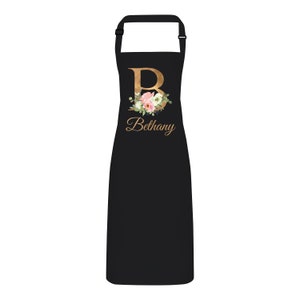 Op de afbeelding: Zwarte schort met een gouden bloemmotief initial "B" en de naam "Bethany" in goud gedrukt.
