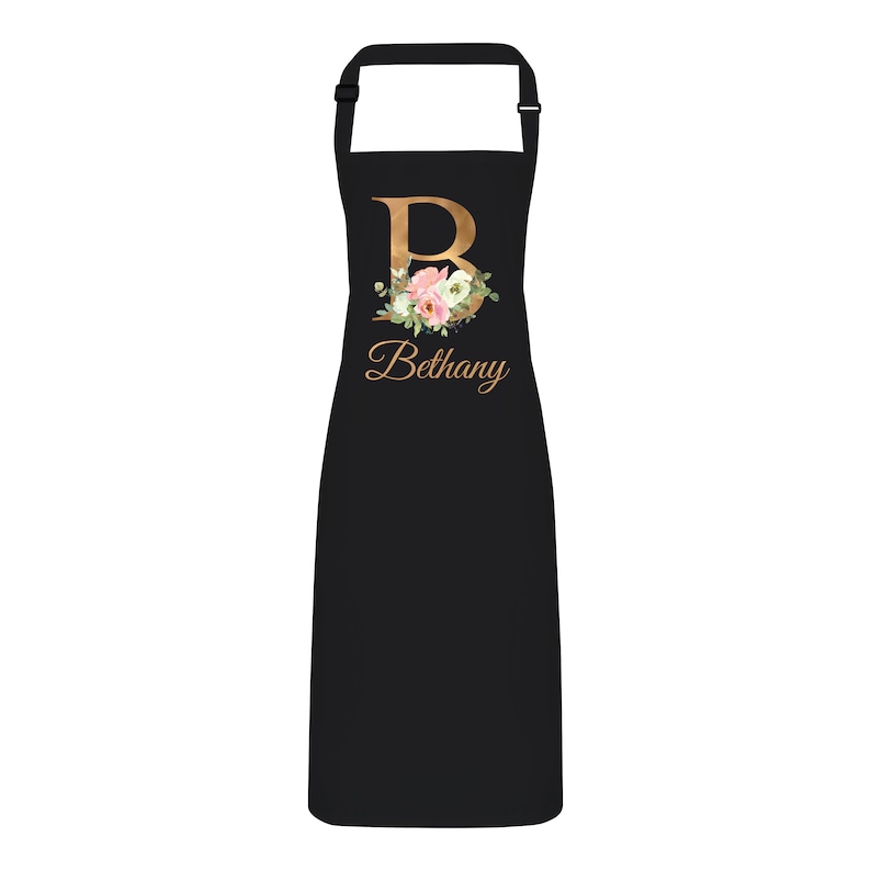 Op de afbeelding: Zwarte schort met een gouden bloemmotief initial "B" en de naam "Bethany" in goud gedrukt.
