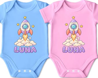 Könnte beinhalten: Zwei kurzärmelige Babybodys, einer in Hellblau und einer in Hellrosa, jeweils mit einer Grafik einer Cartoon-Rakete, die von Wolken abhebt, mit gelben Sternen und dem Namen LUNA in lila Buchstaben mit weißen Sternakzenten bedruckt.
