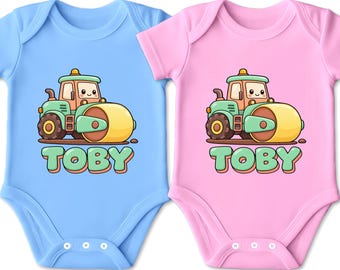 Könnte beinhalten: Zwei kurzärmelige Babybodys, einer in Hellblau und einer in Hellrosa. Beide zeigen eine Cartoon-Illustration einer grünen und gelben Straßenwalze mit einem lächelnden Gesicht über dem Namen TOBY, der in großen, bauchigen grünen Buchstaben geschrieben ist.