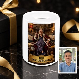 May include: A white ceramic money box featuring a printed image of a person wearing a crown and a red velvet robe while holding a remote control. The text KEN REMOTE CONTROL MONARCH is printed on a gold-coloured plaque at the bottom of the image.