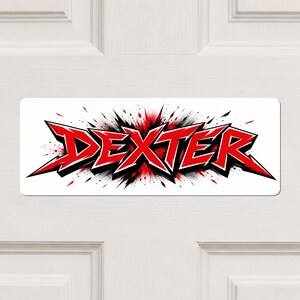 May include: A rectangular sticker featuring the name DEXTER in bold, sharp, red and black lettering. The design includes a dynamic, explosive background effect with red and black paint splatters radiating from behind the text on a white base.