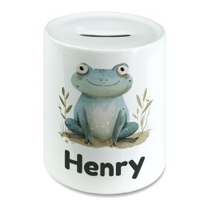 May include: White ceramic piggy bank with a blue frog illustration and the name "Henry" printed on the front.