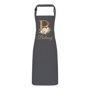 Op de afbeelding: Een grijze schort met een gouden bloemenontwerp en de letter "B" met de naam "Bethany" eronder.