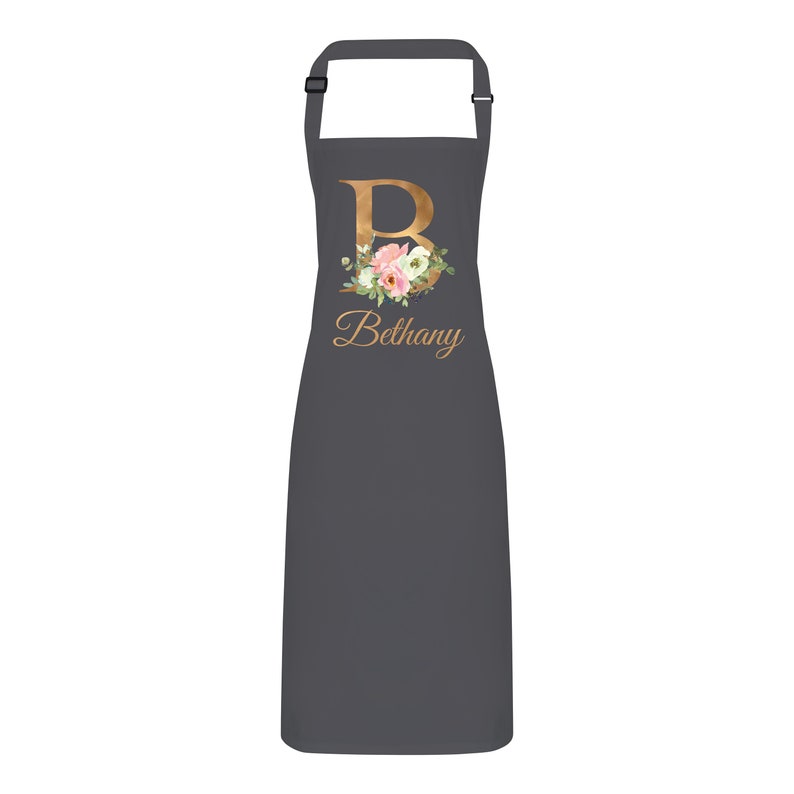 Op de afbeelding: Een grijze schort met een gouden bloemenontwerp en de letter "B" met de naam "Bethany" eronder.