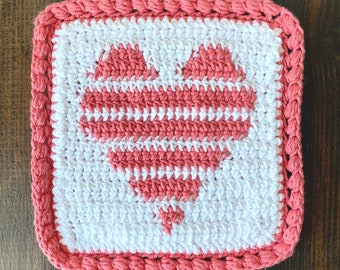 May include: A square crochet coaster featuring a white background with a central heart motif created in alternating horizontal stripes of coral pink and white yarn. The square piece is finished with a textured coral pink border around the edge.