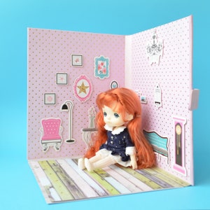 Könnte beinhalten: Eine Puppe mit roten Haaren und blauen Augen sitzt in einem rosa Puppenhaus. Das Puppenhaus hat eine rosa gepunktete Wand mit Aufklebern von Möbeln und Kunstwerken. Der Boden besteht aus Holzoptik-Papier.