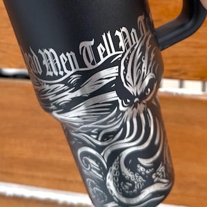 May include: Black stainless steel travel mug with a black handle and a black lid. The mug has a silver graphic of an octopus with the text "Old Men Tell No Tales" printed on it. The mug is made by Stanley.