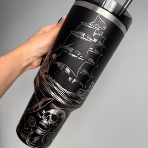 K&ouml;nnte beinhalten: Schwarzer Edelstahl-Reisebecher mit einem lasergravierten Design eines Piratenschiffs, eines Sch&auml;dels und von M&uuml;nzen. Der Becher hat einen Deckel mit einem Strohhalm.