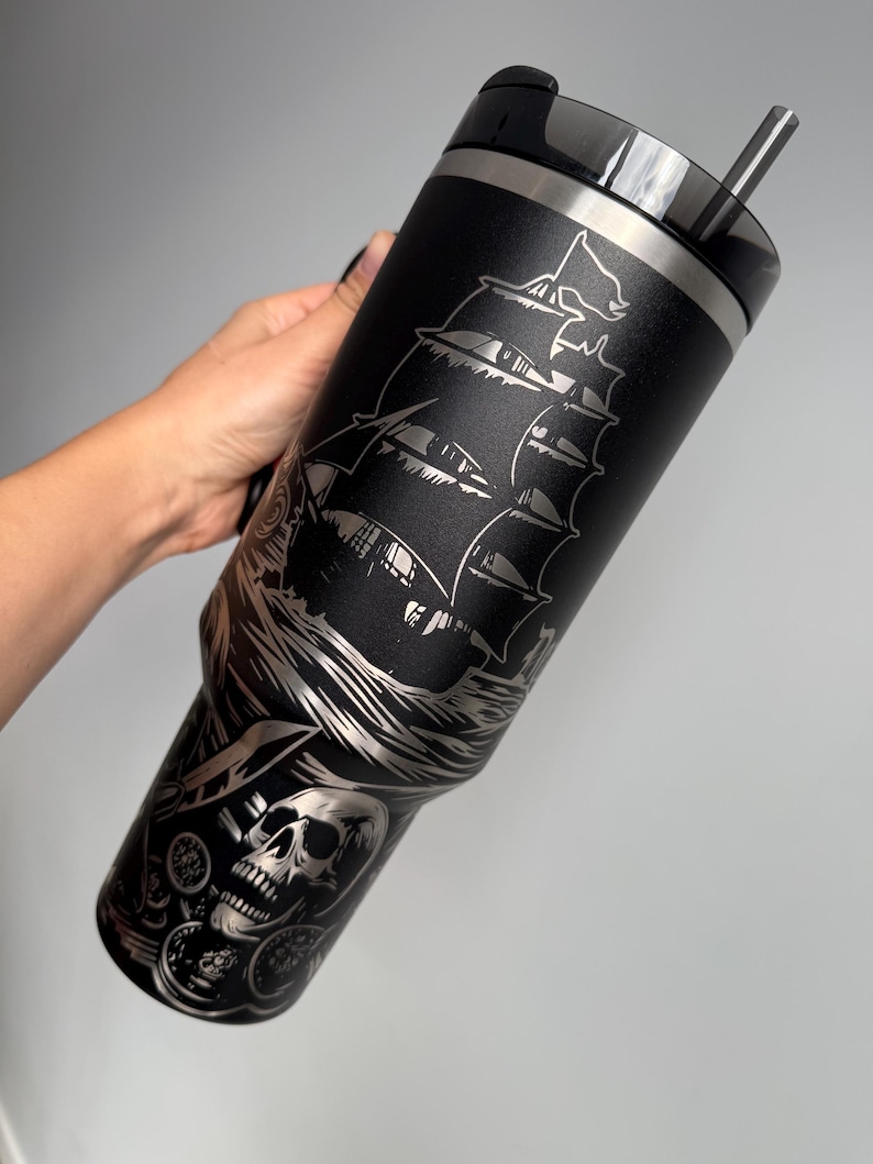 K&ouml;nnte beinhalten: Schwarzer Edelstahl-Reisebecher mit einem lasergravierten Design eines Piratenschiffs, eines Sch&auml;dels und von M&uuml;nzen. Der Becher hat einen Deckel mit einem Strohhalm.