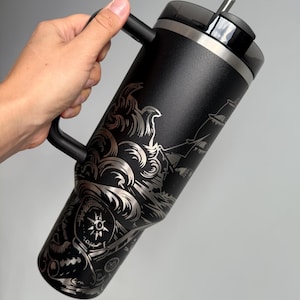 K&ouml;nnte beinhalten: Schwarze Reise-Thermoskanne aus Edelstahl mit schwarzem Griff und einem Deckel mit Strohhalm. Die Thermoskanne ist mit einem gravierten Design eines Schiffes, das durch Wellen f&auml;hrt, mit einem Kompass und anderen nautischen Elementen verziert.