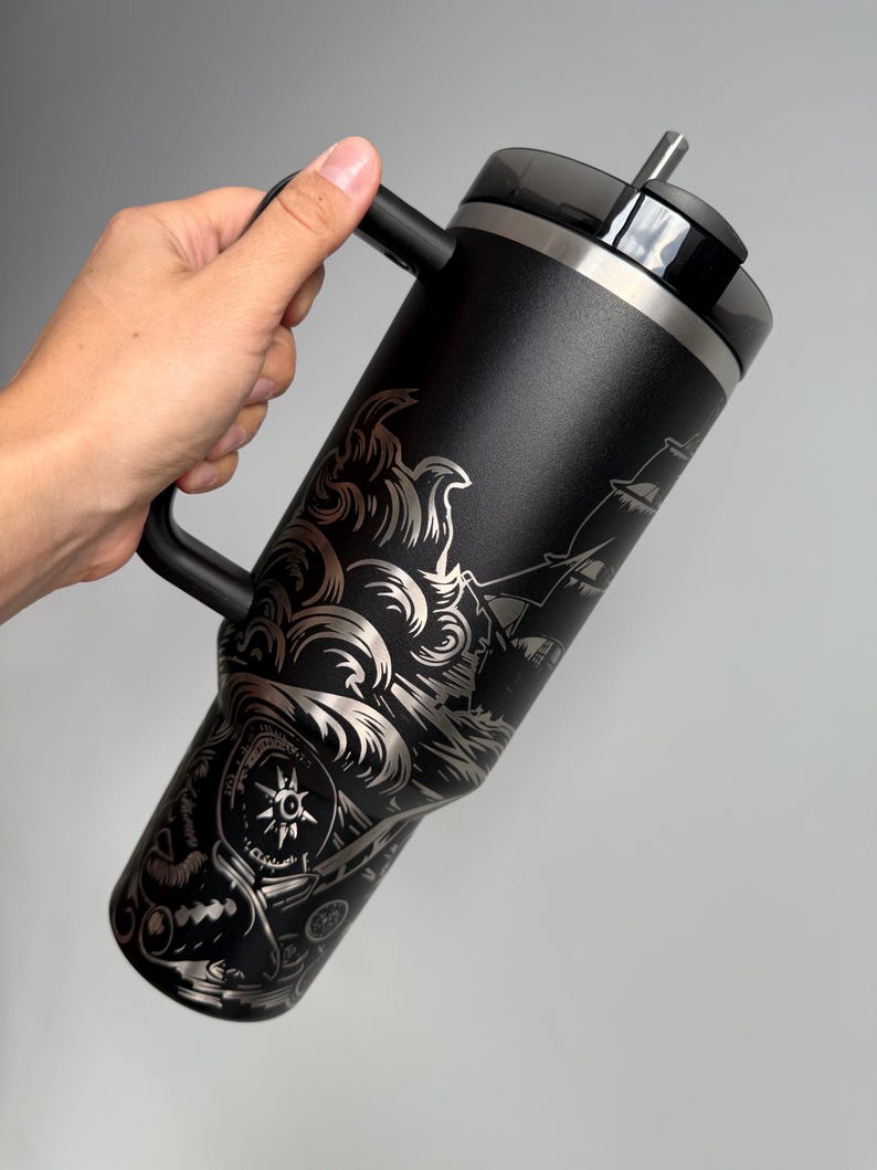 K&ouml;nnte beinhalten: Schwarze Reise-Thermoskanne aus Edelstahl mit schwarzem Griff und einem Deckel mit Strohhalm. Die Thermoskanne ist mit einem gravierten Design eines Schiffes, das durch Wellen f&auml;hrt, mit einem Kompass und anderen nautischen Elementen verziert.