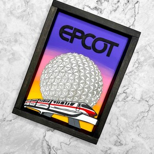 May include: Framed artwork with the word "EPCOT" above a geometric sphere and a monorail train. The background transitions from purple to yellow. The frame is black.
