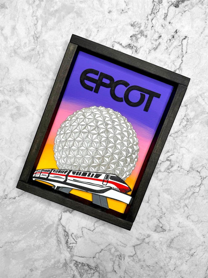 May include: Framed artwork with the word "EPCOT" above a geometric sphere and a monorail train. The background transitions from purple to yellow. The frame is black.