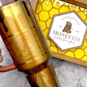 May include: A gold-colored tumbler with a honey-themed design, featuring dripping honey and honeycomb patterns. The tumbler has a brown handle and a lid. A wooden tray with a honeycomb design and a sign that reads "Honey Co." is also visible.