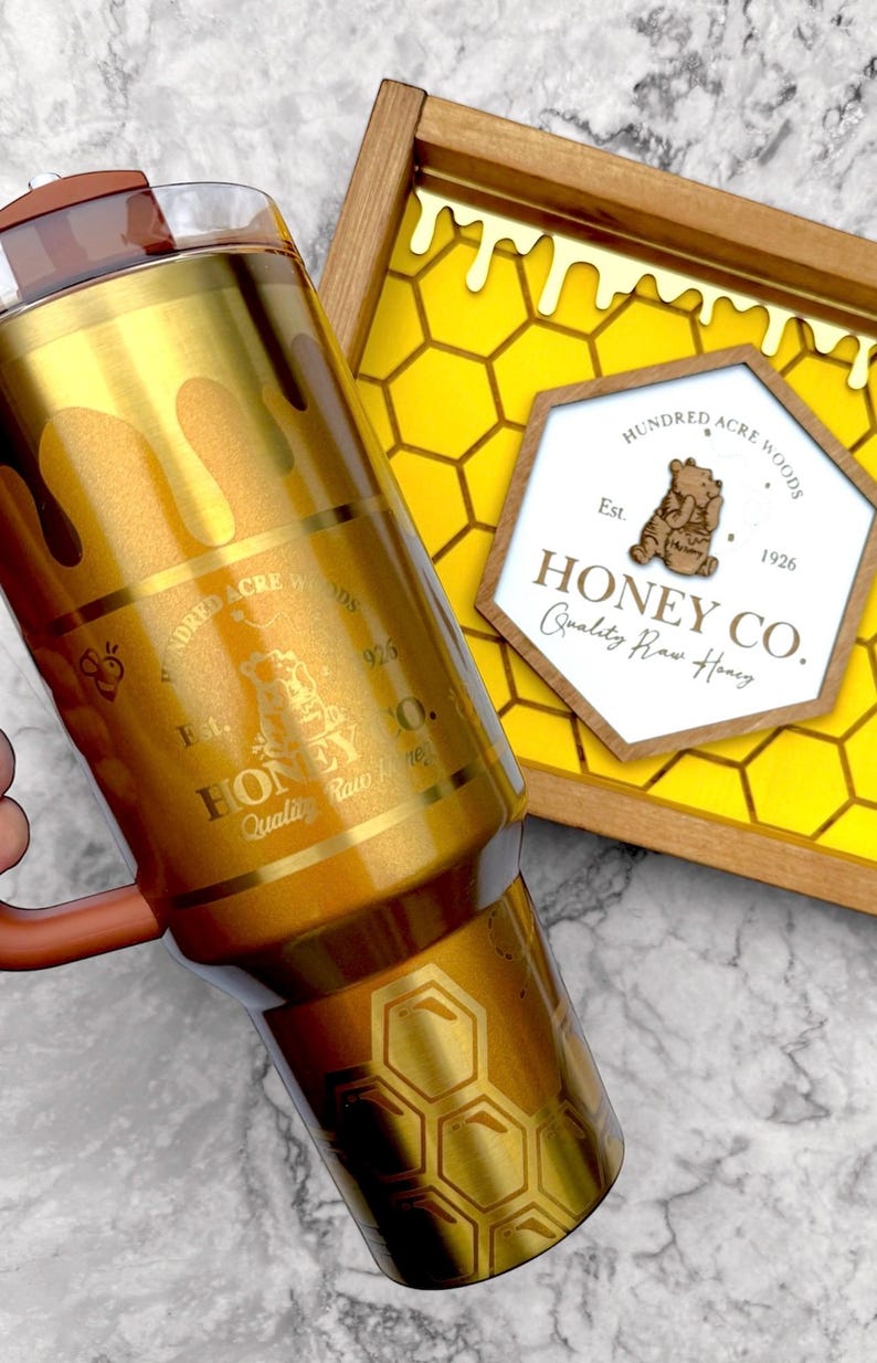 May include: A gold-colored tumbler with a honey-themed design, featuring dripping honey and honeycomb patterns. The tumbler has a brown handle and a lid. A wooden tray with a honeycomb design and a sign that reads "Honey Co." is also visible.