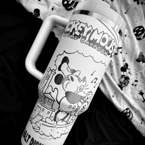 May include: A white travel mug with a large handle, featuring a black and white illustration of Mickey Mouse and the text "Mickey Mouse Sound Cartoon." The mug has a silver band and a white lid. The background is a patterned fabric.