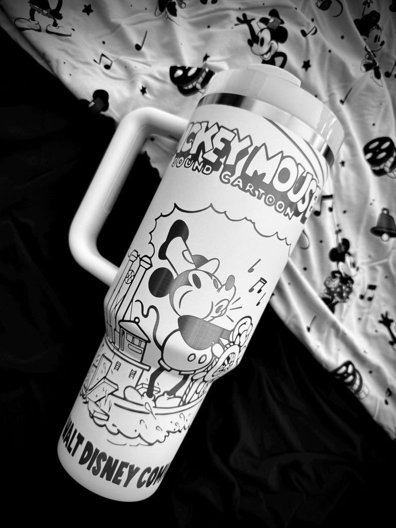May include: A white travel mug with a large handle, featuring a black and white illustration of Mickey Mouse and the text "Mickey Mouse Sound Cartoon." The mug has a silver band and a white lid. The background is a patterned fabric.