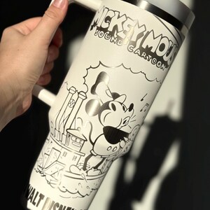 May include: White tumbler with a black and white Mickey Mouse cartoon illustration. The tumbler has a handle, a grey lid, and a straw. The text "Mickey Mouse Sound Cartoon" and "Walt Disney Co" are also visible. The tumbler is likely made of stainless steel.