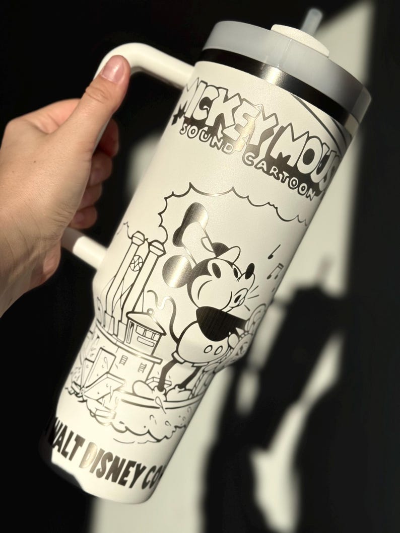 May include: White tumbler with a black and white Mickey Mouse cartoon illustration. The tumbler has a handle, a grey lid, and a straw. The text "Mickey Mouse Sound Cartoon" and "Walt Disney Co" are also visible. The tumbler is likely made of stainless steel.