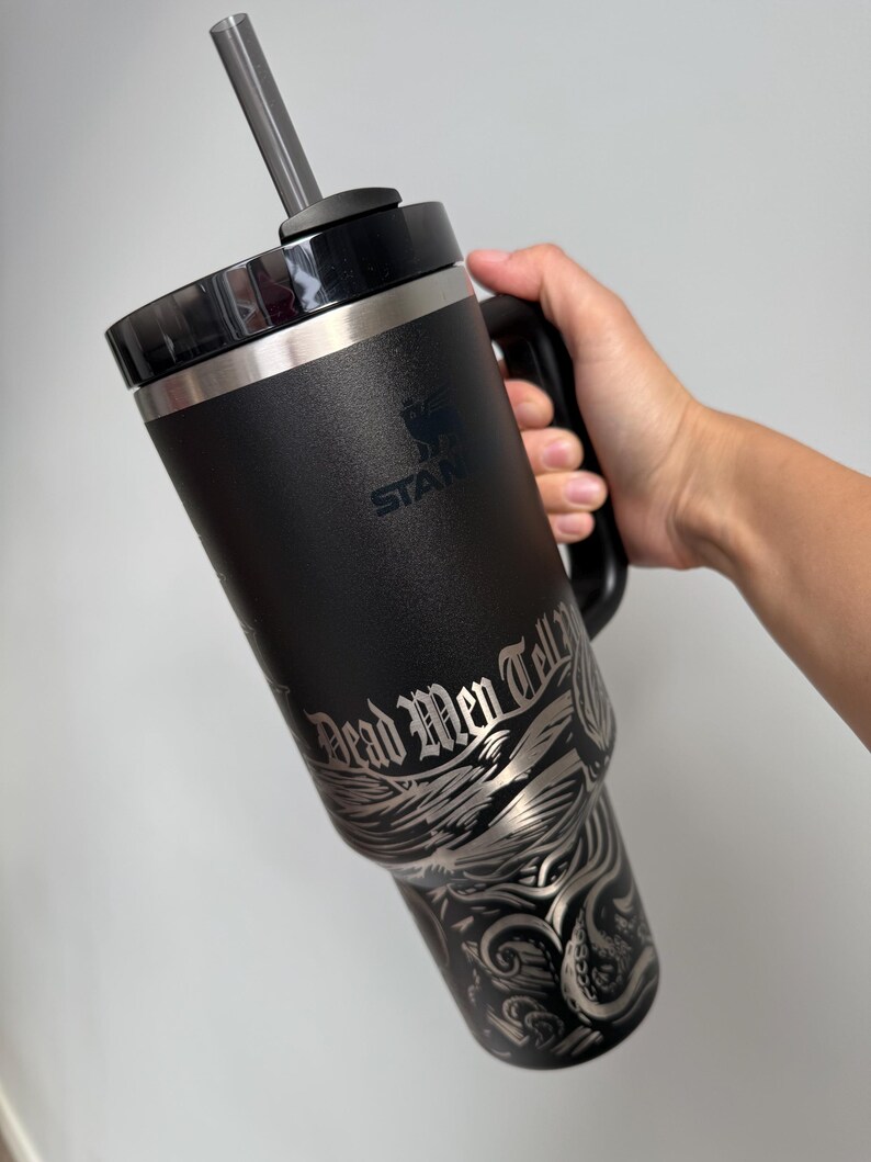 K&ouml;nnte beinhalten: Schwarze Reise-Thermoskanne aus Edelstahl mit schwarzem Griff und klarem Strohhalm. Die Kanne hat ein silbernes Design mit einem Kraken und Wellen mit dem Text "Dead Men Tell No Tales".