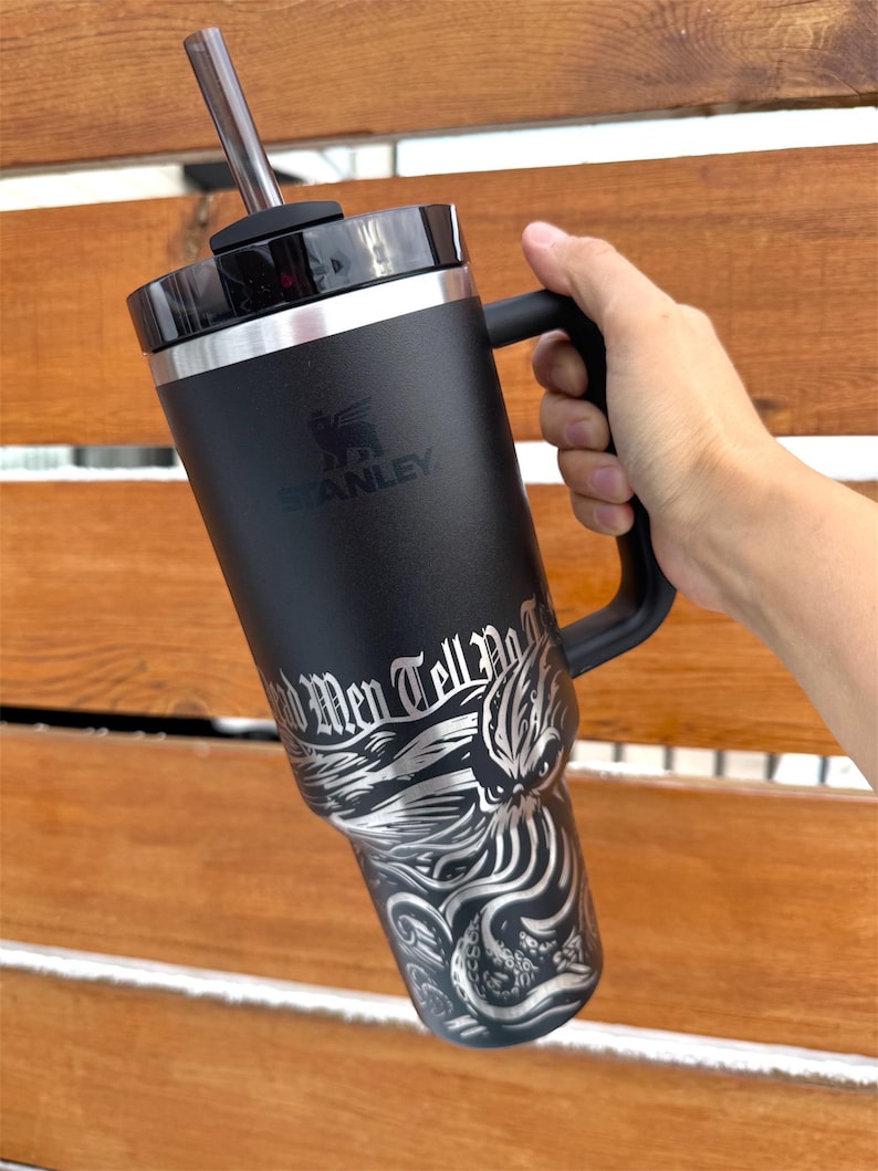 May include: Black stainless steel travel mug with a black handle and a black lid. The mug has a silver graphic of an octopus with the text "Old Men Tell No Tales" printed on it. The mug is made by Stanley.