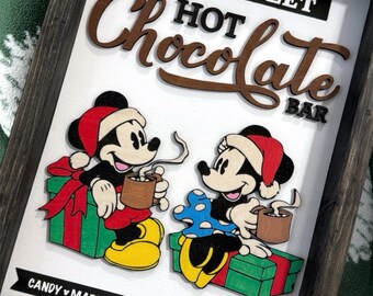 Könnte beinhalten: Holzschild mit dunklem Rahmen, das Mickey und Minnie Mouse mit Weihnachtsmützen zeigt, die Tassen heiße Schokolade halten. Das Schild lautet "Mainstreet Hot Chocolate Bar" mit Text: "Candy, Marshmallow, Cinnamon, Magic."