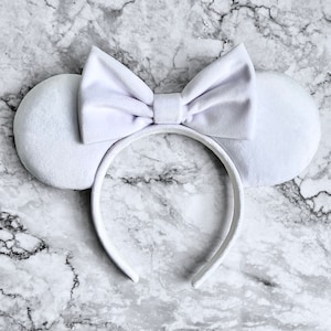May include: A white headband with attached mouse ears and a bow. The ears are round and the bow is centered on the headband. The headband and ears are made of a soft, plush material.