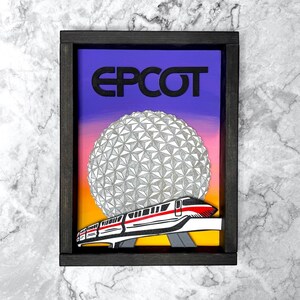 May include: Framed artwork with the word "EPCOT" in black above a geometric sphere and a monorail train. The background fades from purple to orange and yellow. The frame is dark brown.
