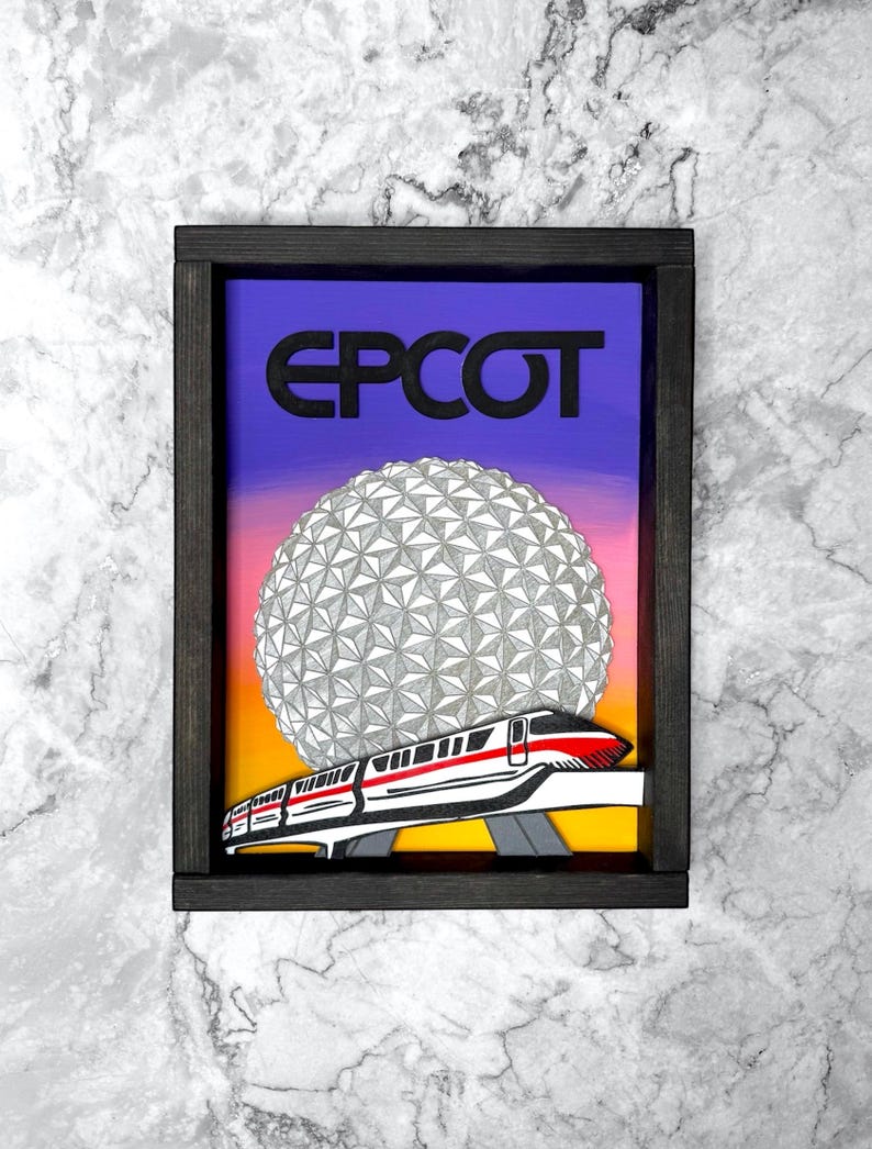 May include: Framed artwork with the word "EPCOT" in black above a geometric sphere and a monorail train. The background fades from purple to orange and yellow. The frame is dark brown.
