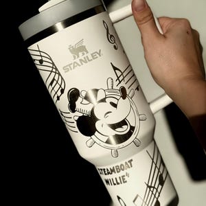 May include: White Stanley travel mug with a grey lid and handle, featuring a black and white Steamboat Willie design. The mug has musical notes and the text "Steamboat Willie". Includes a straw.