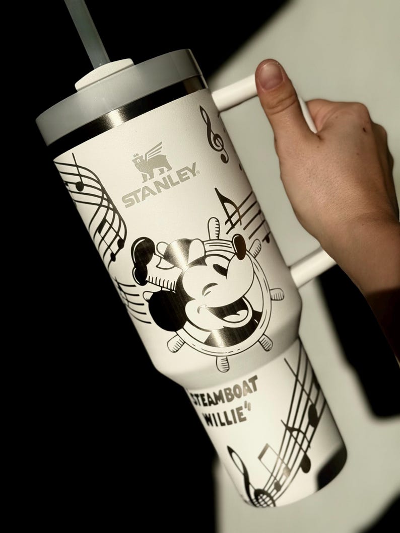 May include: White Stanley travel mug with a grey lid and handle, featuring a black and white Steamboat Willie design. The mug has musical notes and the text "Steamboat Willie". Includes a straw.