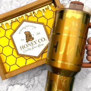 May include: A gold-colored Stanley tumbler with the word "HUNNY" and a bee graphic. The tumbler has a brown handle and lid, and a clear straw. A wooden tray with a honeycomb design and "Honey Co." text is also visible.