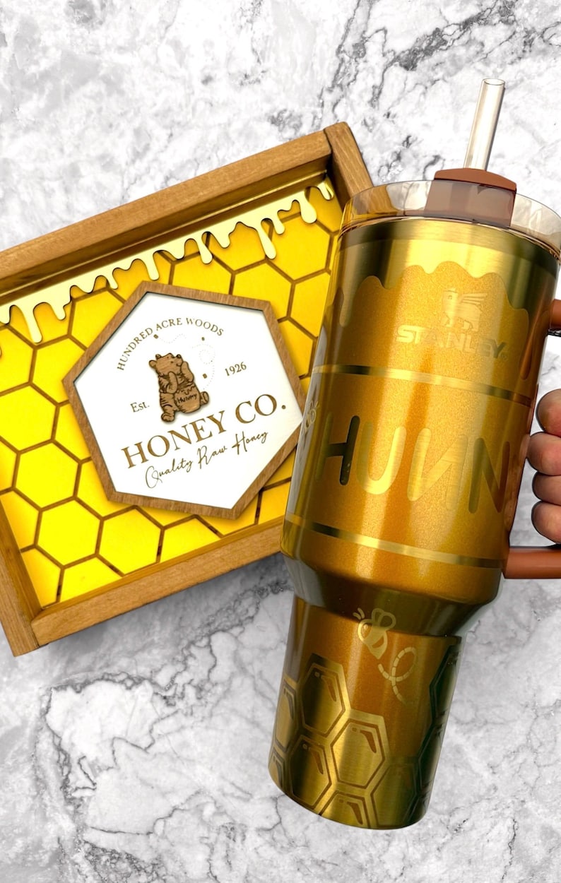 May include: A gold-colored Stanley tumbler with the word "HUNNY" and a bee graphic. The tumbler has a brown handle and lid, and a clear straw. A wooden tray with a honeycomb design and "Honey Co." text is also visible.