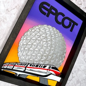 May include: Framed artwork with the word "EPCOT" in black above a geometric sphere and a monorail train. The background transitions from orange to purple. The frame is dark brown.