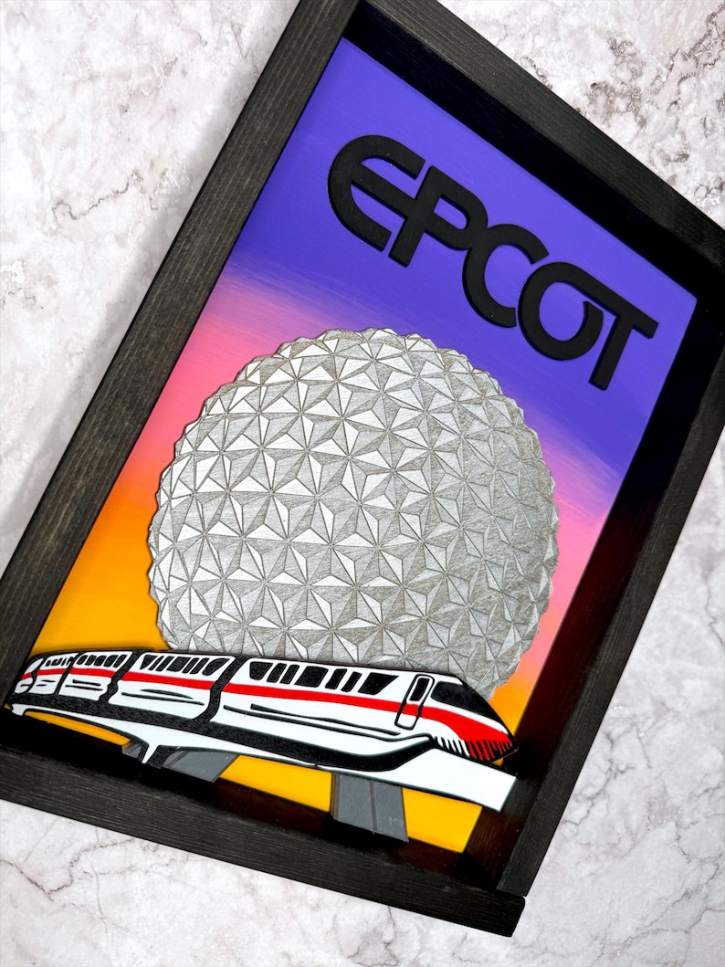 May include: Framed artwork with the word "EPCOT" in black above a geometric sphere and a monorail train. The background transitions from orange to purple. The frame is dark brown.