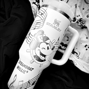 May include: White Stanley tumbler with a Steamboat Willie design and musical notes. The tumbler has a handle and a straw. The Stanley logo is visible. The design includes a smiling Mickey Mouse character at the helm of a ship.
