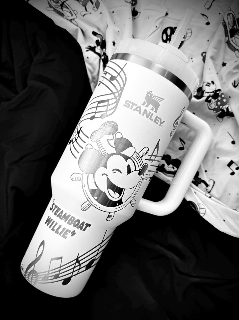 May include: White Stanley tumbler with a Steamboat Willie design and musical notes. The tumbler has a handle and a straw. The Stanley logo is visible. The design includes a smiling Mickey Mouse character at the helm of a ship.