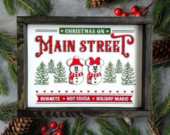 Może przedstawiać: Oprawiony drewniany znak z napisem "Christmas on Main Street" w kolorze czerwonym i zielonym. Znak przedstawia postacie bałwanków i słowa "Beignets, Hot Cocoa, Holiday Magic". Rama jest ciemnobrązowa.