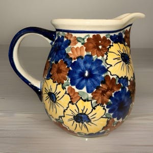 May include: A ceramic pitcher with a white handle and spout, and a blue handle. The pitcher is decorated with a floral pattern of yellow, blue, and brown flowers. The rim is white with a blue stripe.