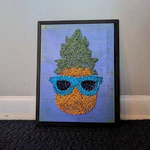 May include: A string art pineapple with a blue and green leaf and blue sunglasses on a purple background.