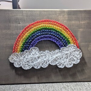 May include: A rainbow made of colorful yarn is strung on nails and attached to a wooden board. A white yarn cloud is below the rainbow.